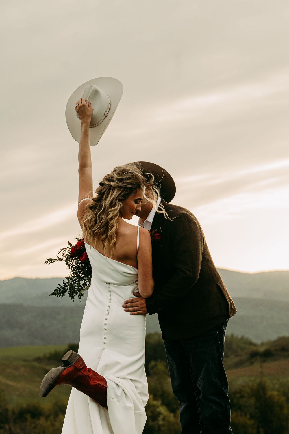 Bride in white gown and red boots raises hat while kissing groom in brown hat at sunset mountain wedding.