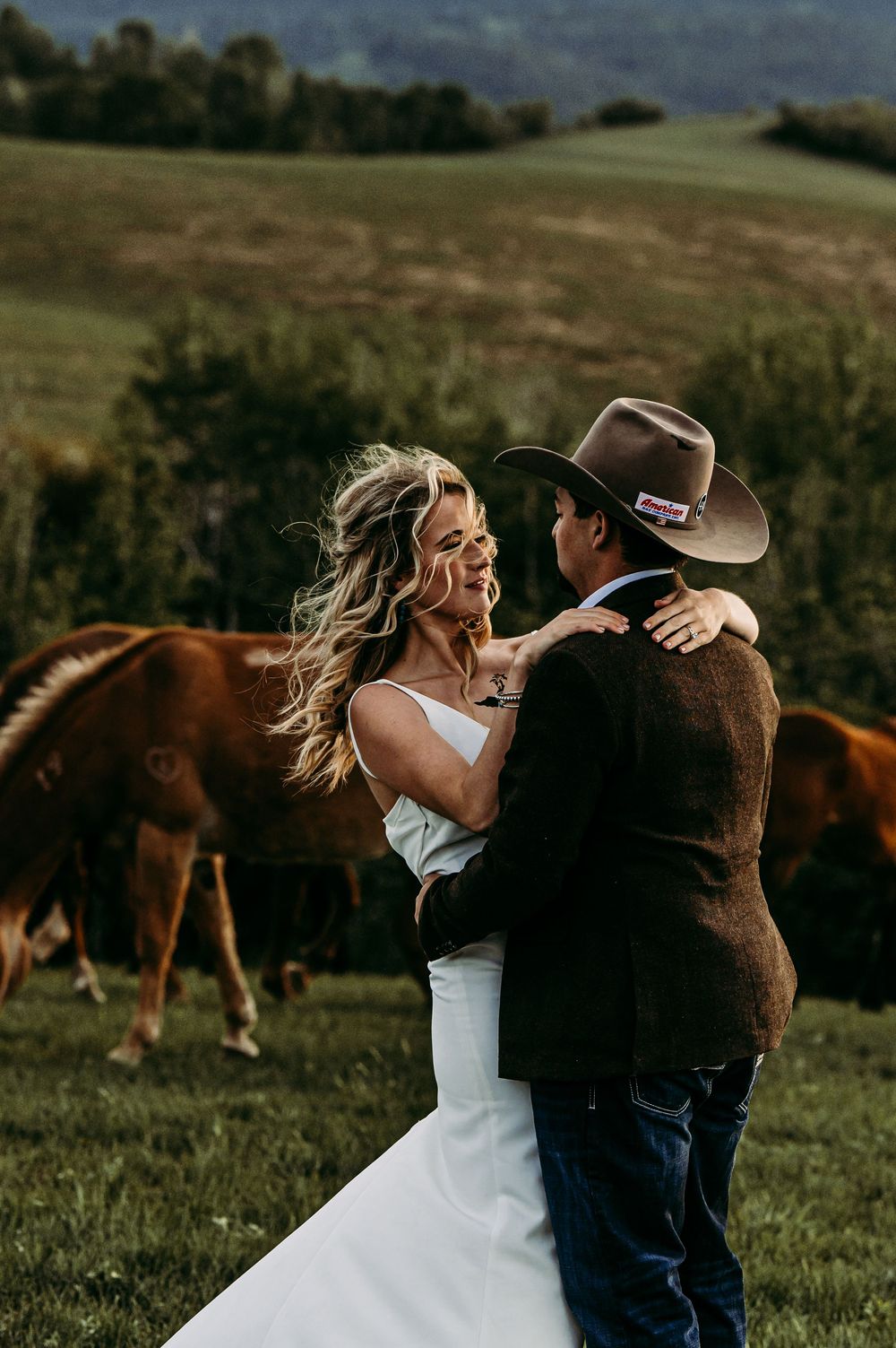 A bride and groom share a romantic dance in a green pasture with horses grazing behind them on their country wedding day.