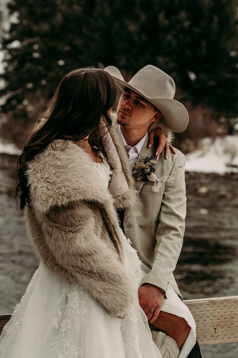 Bride in fur wrap and groom in tan cowboy hat share a romantic winter wedding moment by a snowy river.