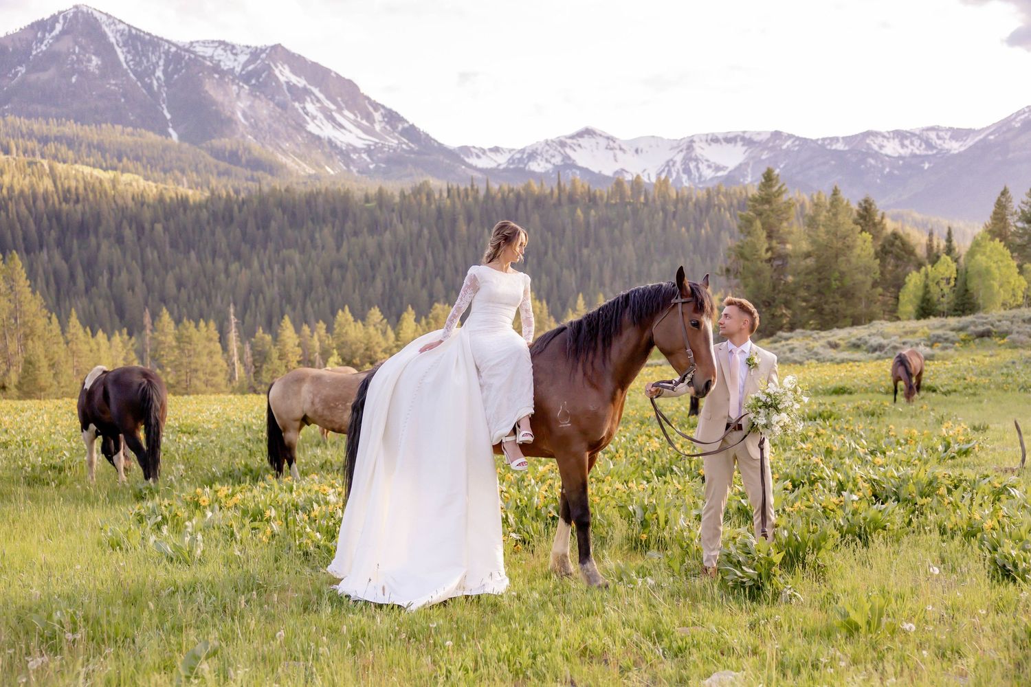 A bride in a white gown sits on a horse with a groom beside her in a mountain meadow in Jackson, Wyoming.