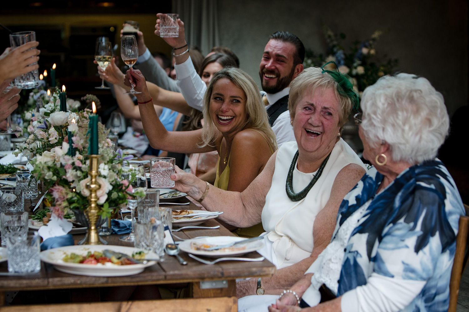 Joyful wedding guests laughing and toasting with champagne glasses at an elegant floral dinner table celebration.