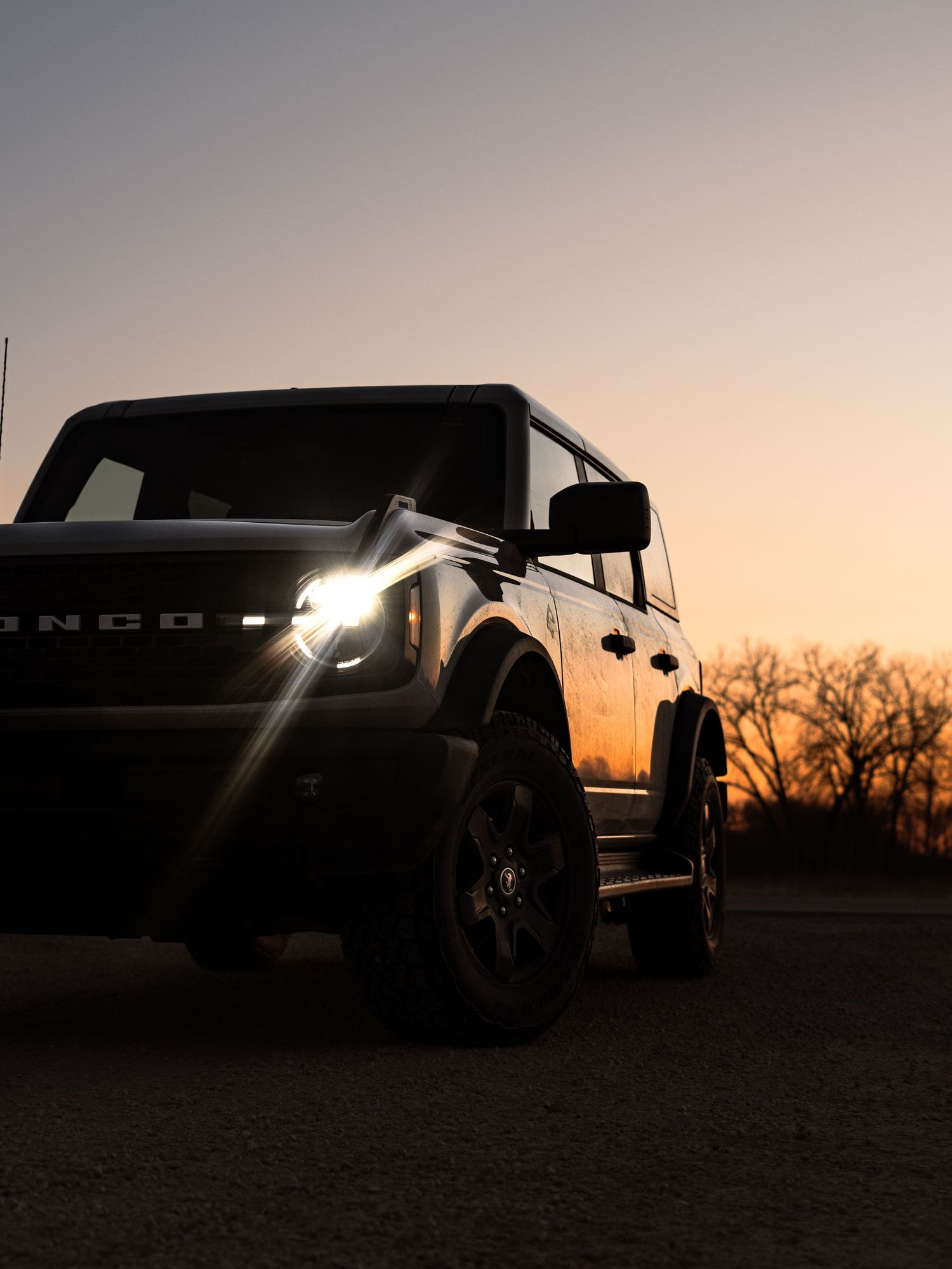 Ford Bronco SUV silhouetted against a dramatic sunset sky with headlights glowing and bare trees in the background.