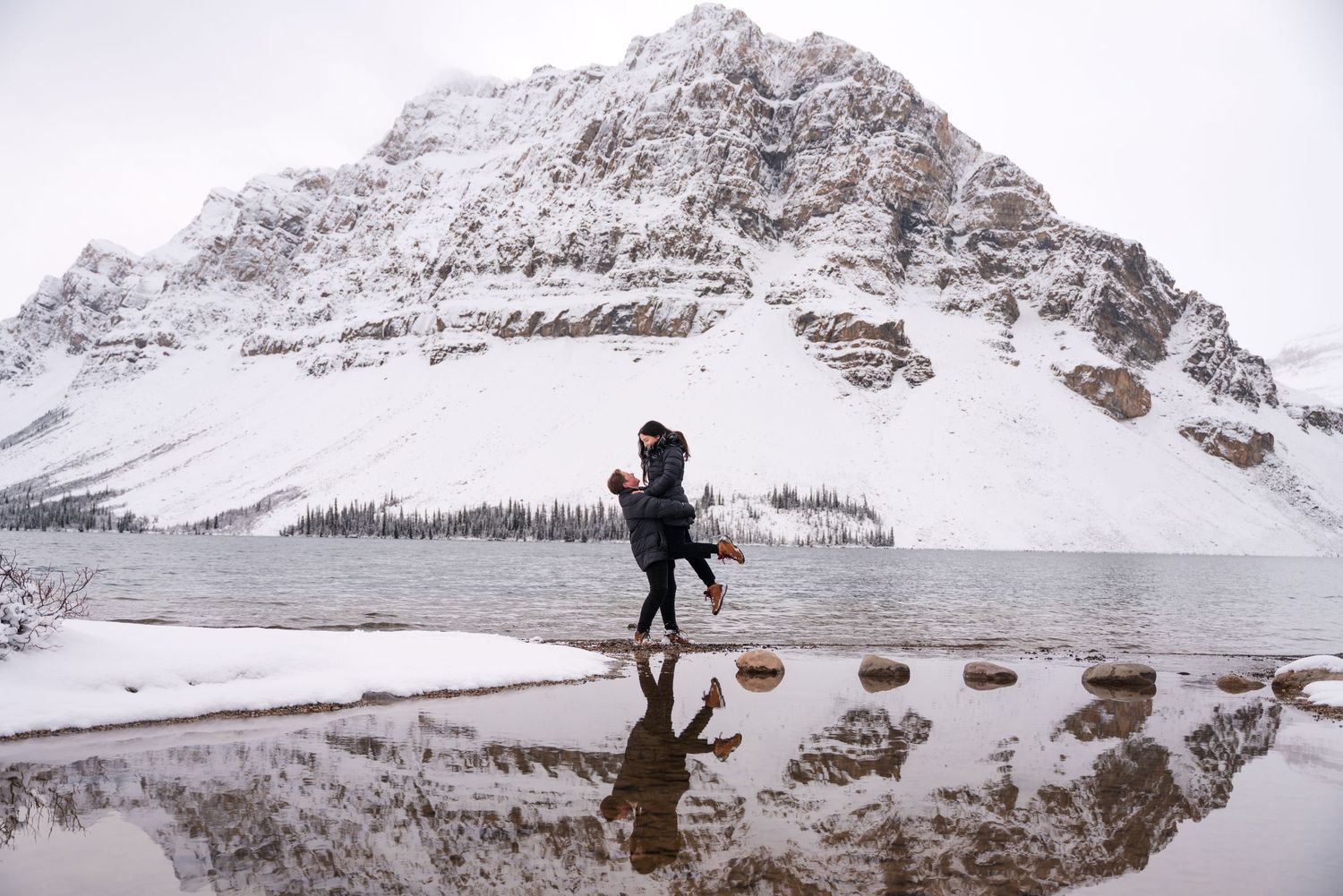 Two women embracing by Bow Lake, Banff Alberta a snowy mountain lake with their reflection visible in the calm water below.