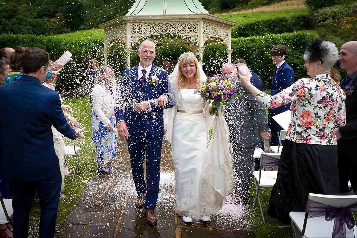 Bride and groom walk through confetti shower with smiling guests at outdoor garden wedding ceremony.