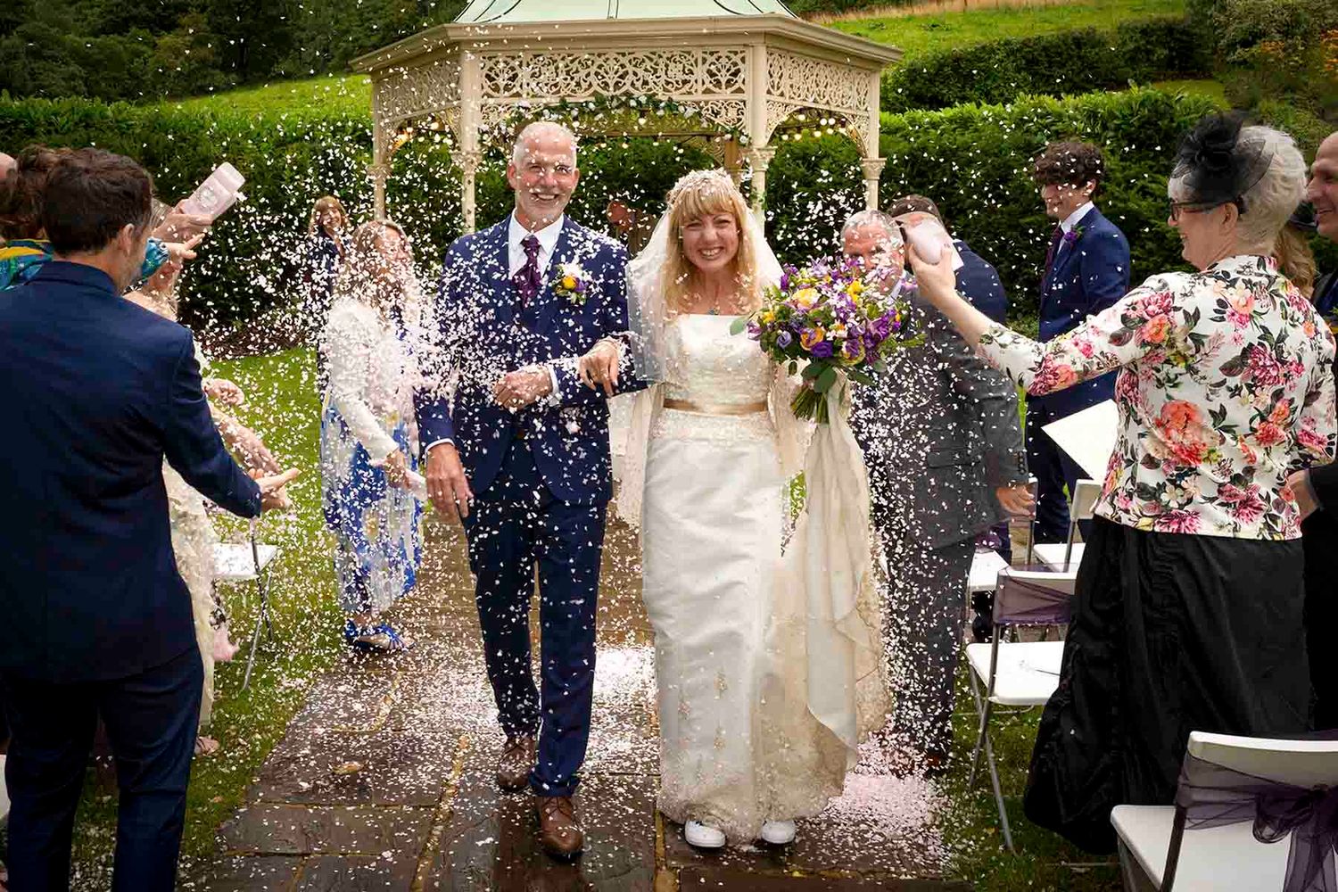 Bride and groom walk through confetti shower with smiling guests at outdoor garden wedding ceremony.