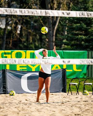 Oregon Beach Volleyball player serves ball on sandy court with green and yellow Oregon Ducks banners in background.