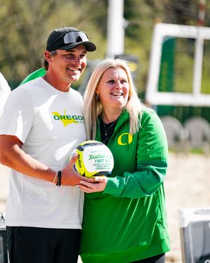 Two people in Oregon Ducks gear smile while holding a volleyball on a sunny beach volleyball court.