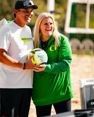 Two Oregon Ducks staff members smile while holding a volleyball on a beach volleyball court.