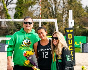 Oregon Ducks beach volleyball player #12 poses with two supporters wearing green Oregon Ducks gear, holding flowers on a sunny day.