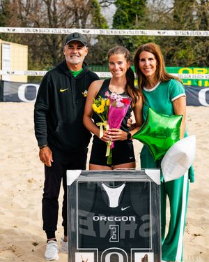 Oregon volleyball player honored on senior day with family, holding flowers and jersey on beach volleyball court.