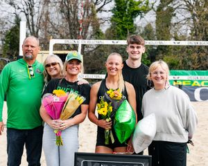 Beach volleyball players celebrate with family, holding flowers and a trophy near a sand court outdoors.