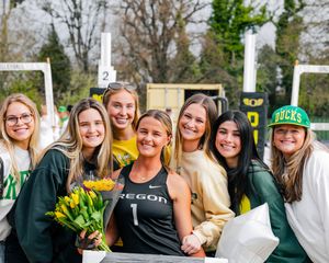 Group of young women posing together outdoors at a track event, one holding yellow tulips wearing an Oregon jersey.
