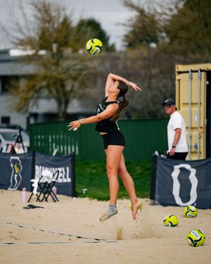 Female beach volleyball player jumps to serve yellow ball on sand court during outdoor match.