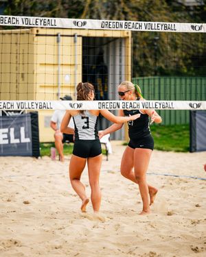 Two Oregon Beach Volleyball players celebrate on sand court, wearing black and white uniforms, with net and signage behind them.