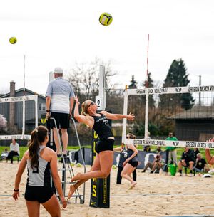 Female beach volleyball players compete outdoors on sand court, one player jumping to spike ball over net.