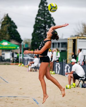 Female beach volleyball player leaps to serve a yellow ball on sandy court during outdoor tournament.