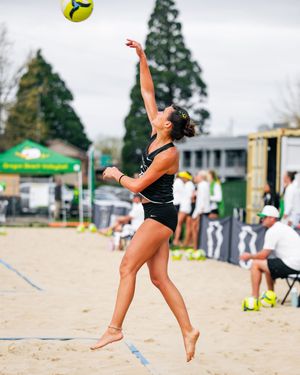 Female beach volleyball player leaping to serve ball on sandy court during outdoor tournament.
