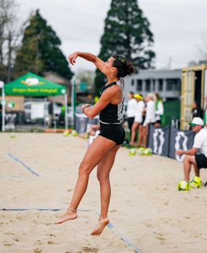 Female beach volleyball player jumping to serve on sandy court with green Oregon Ducks branding visible in background.