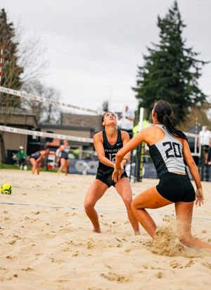 Two beach volleyball players in black uniforms compete on sand court, one setting ball near the net.