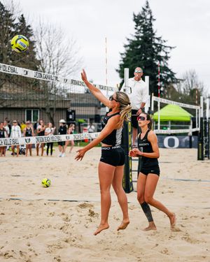 Two female beach volleyball players compete on sand court, one jumping to spike as teammate watches during outdoor tournament.