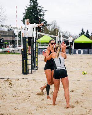 Two beach volleyball players celebrate on sand court during a match, with a referee on elevated stand behind them.