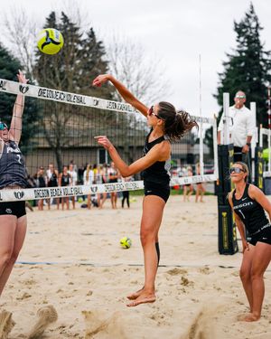 Oregon beach volleyball players compete in a sand court match, with one player jumping to spike the ball over the net.