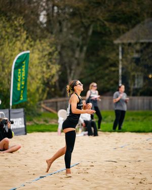 Woman in black athletic wear plays beach volleyball on sand court with a green Beach Volleyball banner in background.