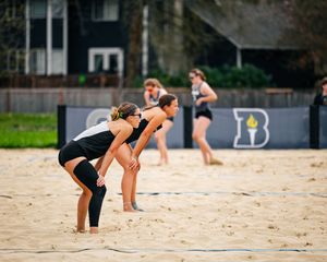 Female beach volleyball players in black uniforms ready for play on sandy court with B logo banner in background.