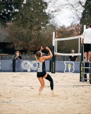 Beach volleyball player in black uniform serving the ball on a sandy court with a net and referee stand visible.