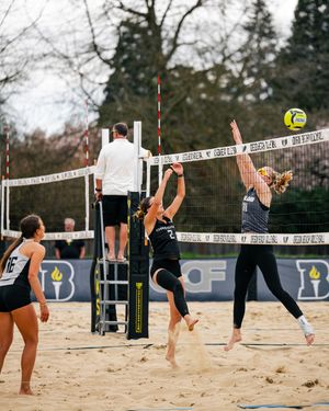 Beach volleyball players compete at the net during an outdoor sand volleyball match with a referee watching.