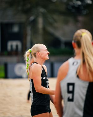 Two female beach volleyball players in black Oregon uniforms smile on a sandy court during a competitive match.