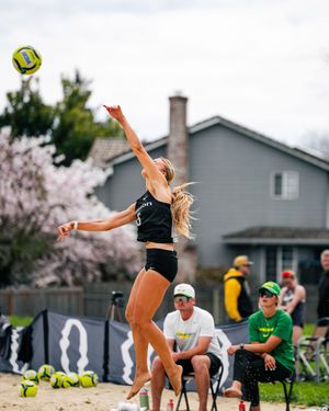 Female beach volleyball player leaping to spike ball mid-air on sand court with cherry blossoms and spectators in background.
