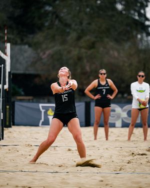 Player #15 in black uniform sets a volleyball on a sandy beach volleyball court during a competitive outdoor match.