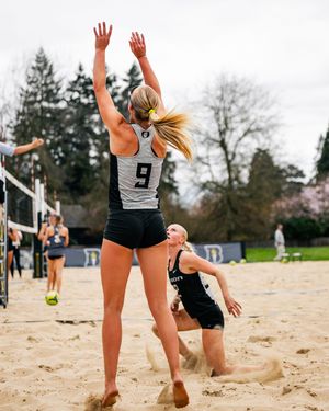 Two female beach volleyball players compete on sand court, player #9 jumping to block at the net.
