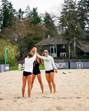 Three beach volleyball players in black uniforms celebrate with high-fives on a sandy court surrounded by trees.