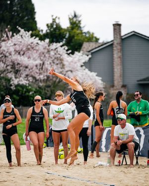 Female beach volleyball player jumping to spike ball in sand court, teammates and coaches watching nearby.