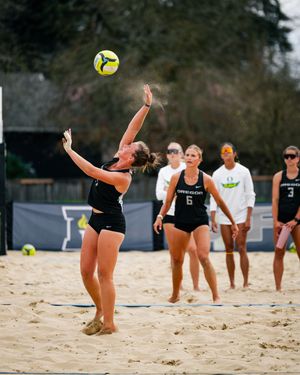 Female beach volleyball player jumping to spike ball during collegiate match on sand court with teammates watching.