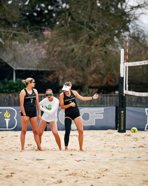 Three beach volleyball players in Oregon Ducks uniforms strategize near the net on a sandy court.