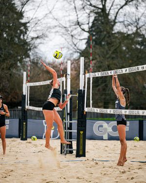 Two women playing beach volleyball, one spiking the ball over the net while the other prepares to block on a sandy court.