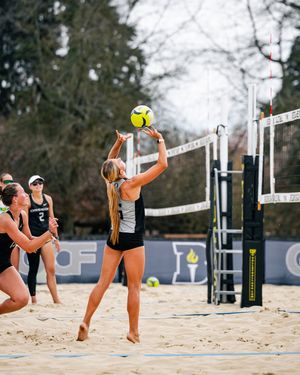 Female beach volleyball player sets ball overhead near net on sandy court with teammates and trees in background.