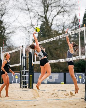 Women playing beach volleyball, one player spiking ball over net while opponent blocks during outdoor sand court match.