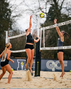 Women playing beach volleyball, one player spiking ball over net while opponent jumps to block on sandy court.