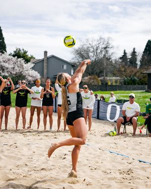 Female beach volleyball player jumps to spike a yellow ball during a sunny outdoor match with teammates watching nearby.