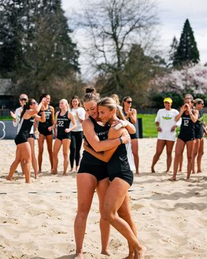 Two Oregon Ducks beach volleyball players embrace on sand court while teammates applaud in background during senior day ceremony.