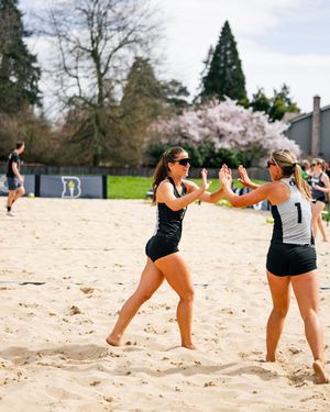 Two beach volleyball players high-five on a sandy court during a sunny spring match, with cherry blossoms blooming in the background.