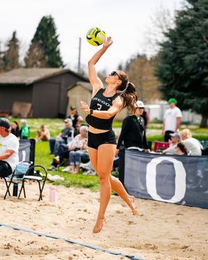 Oregon beach volleyball player jumps to spike a yellow ball during a sunny outdoor match on a sand court.