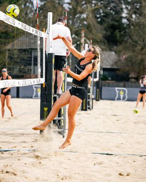 Female beach volleyball player in black Nike outfit jumping to spike ball at the net on a sandy court.