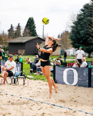 Female beach volleyball player in black uniform jumping to spike a yellow ball on a sandy court.