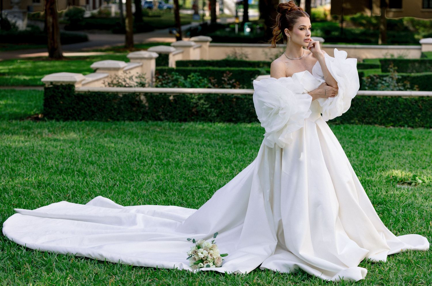 Bride in elegant white off-shoulder ball gown with long train poses on lush green lawn in outdoor garden setting.