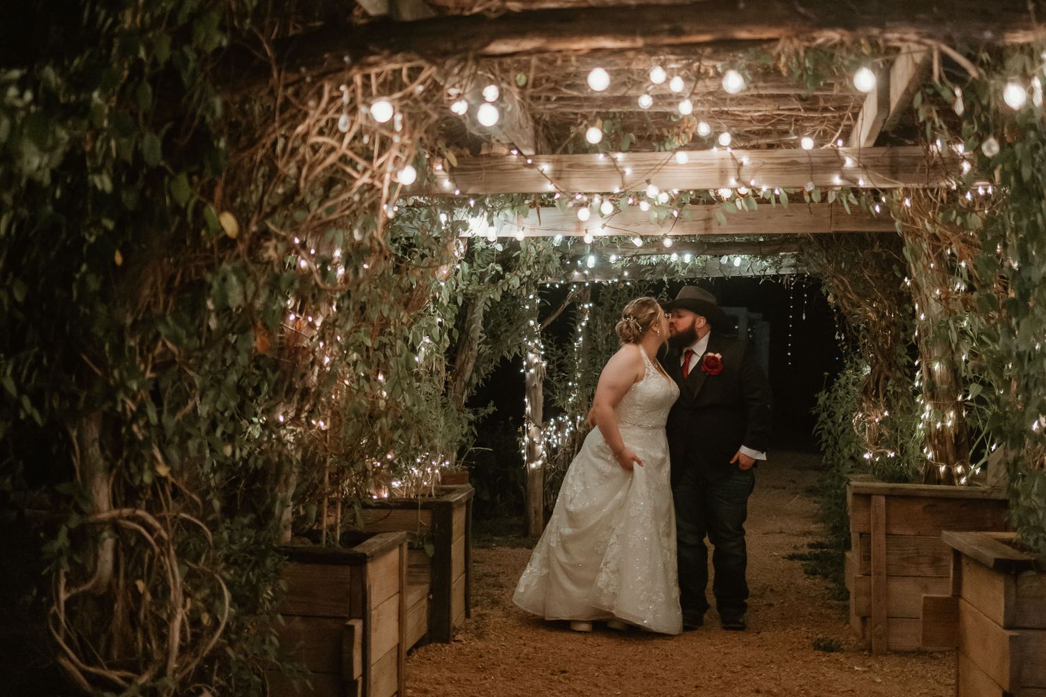 Bride and groom sharing a romantic kiss under a fairy light covered pergola at night.